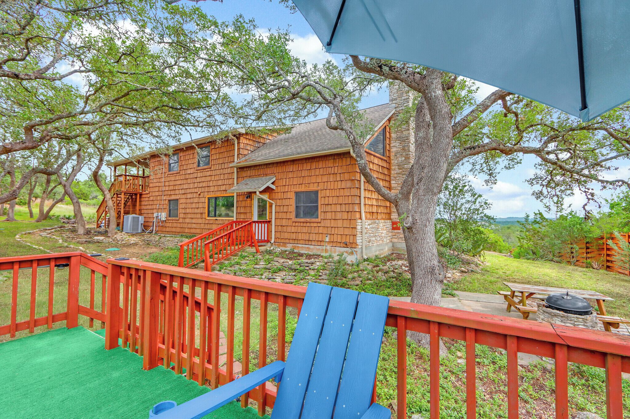 A wooden house with a red deck, a picnic table, and a tree in the background.