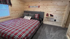 Un dormitorio con una cama, una almohada con la inscripción 'CABIN sweet cabin', un cartel que dice 'NO WAKE ZONE' y un estante con objetos.