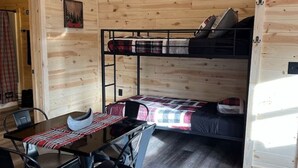 Interior de una cabaña de madera con literas, una mesa de comedor con sillas y una pequeña cocina.