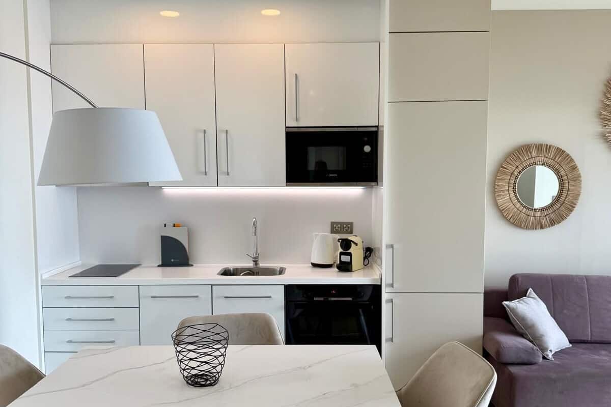 A modern kitchen with white cabinets, a black microwave, and a coffee machine. There is a dining table with chairs and a decorative bowl on it. A sofa and a mirror are visible in the background.