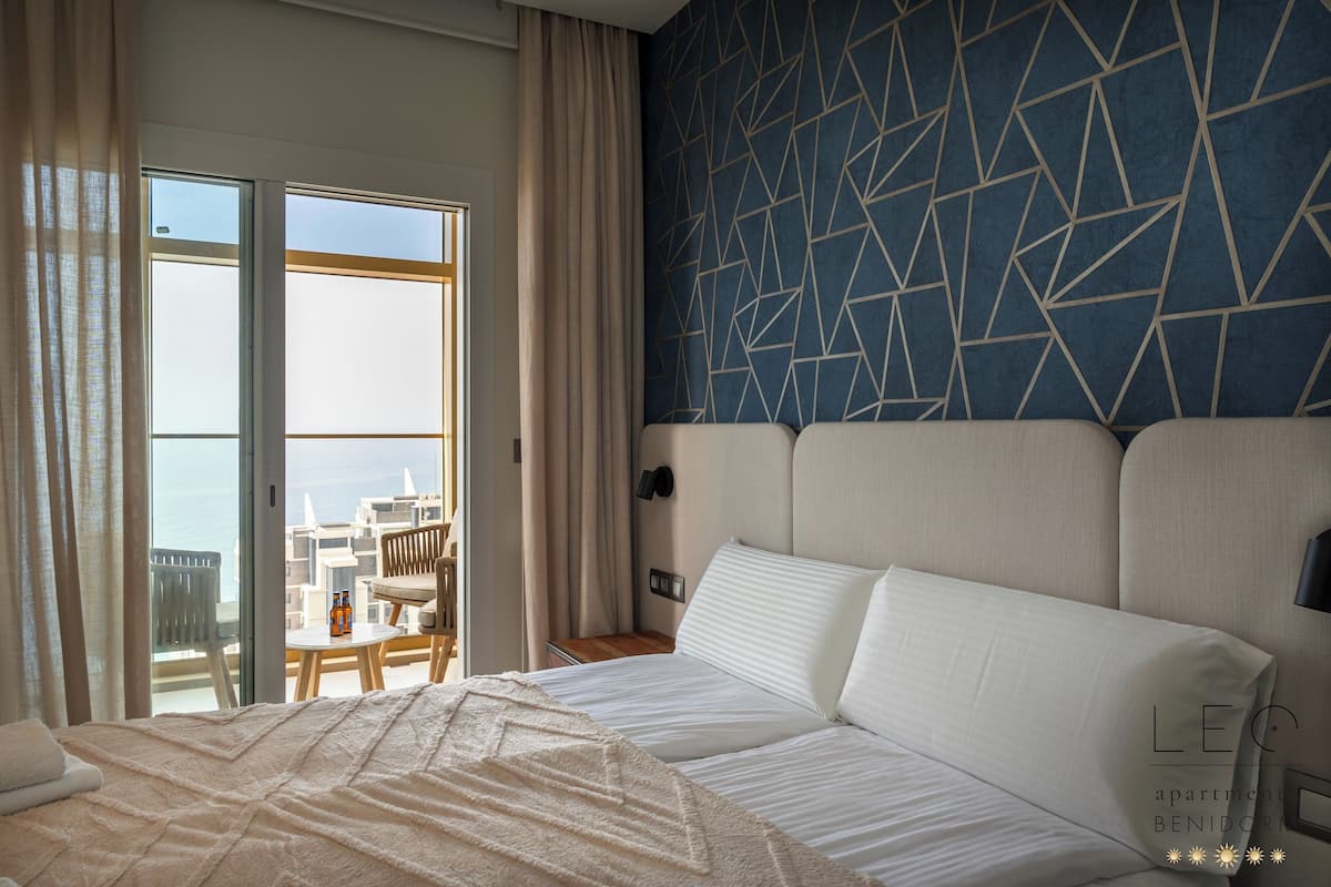 A hotel room with a bed, a view of the sea, a geometric patterned wall, and a balcony with outdoor seating.