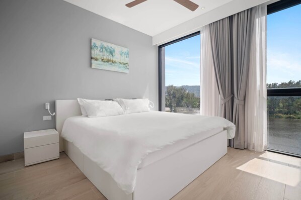 Une chambre moderne avec un grand lit, une table de chevet, un tableau au mur et une vue sur l’extérieur grâce à une grande fenêtre.