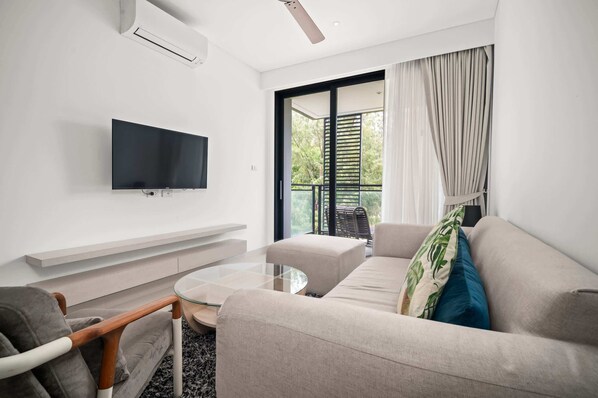 A modern living room with a flat-screen TV, a sofa, a glass coffee table, and a balcony with curtains.
