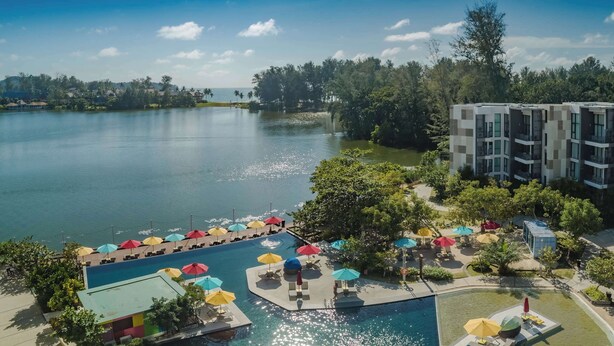 A hotel with a pool area, outdoor umbrellas, and a view of a lake. - Cassia Residences, One Bedroom A Ground Floor (Choeng Thale)