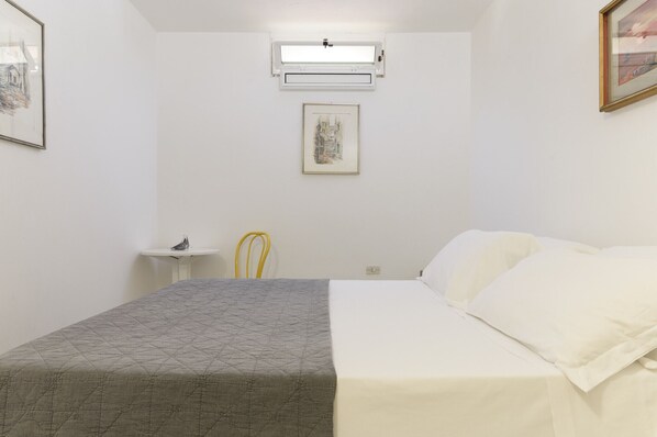 Un dormitorio con cama, una mesita, una silla, un aire acondicionado y un cuadro en la pared.