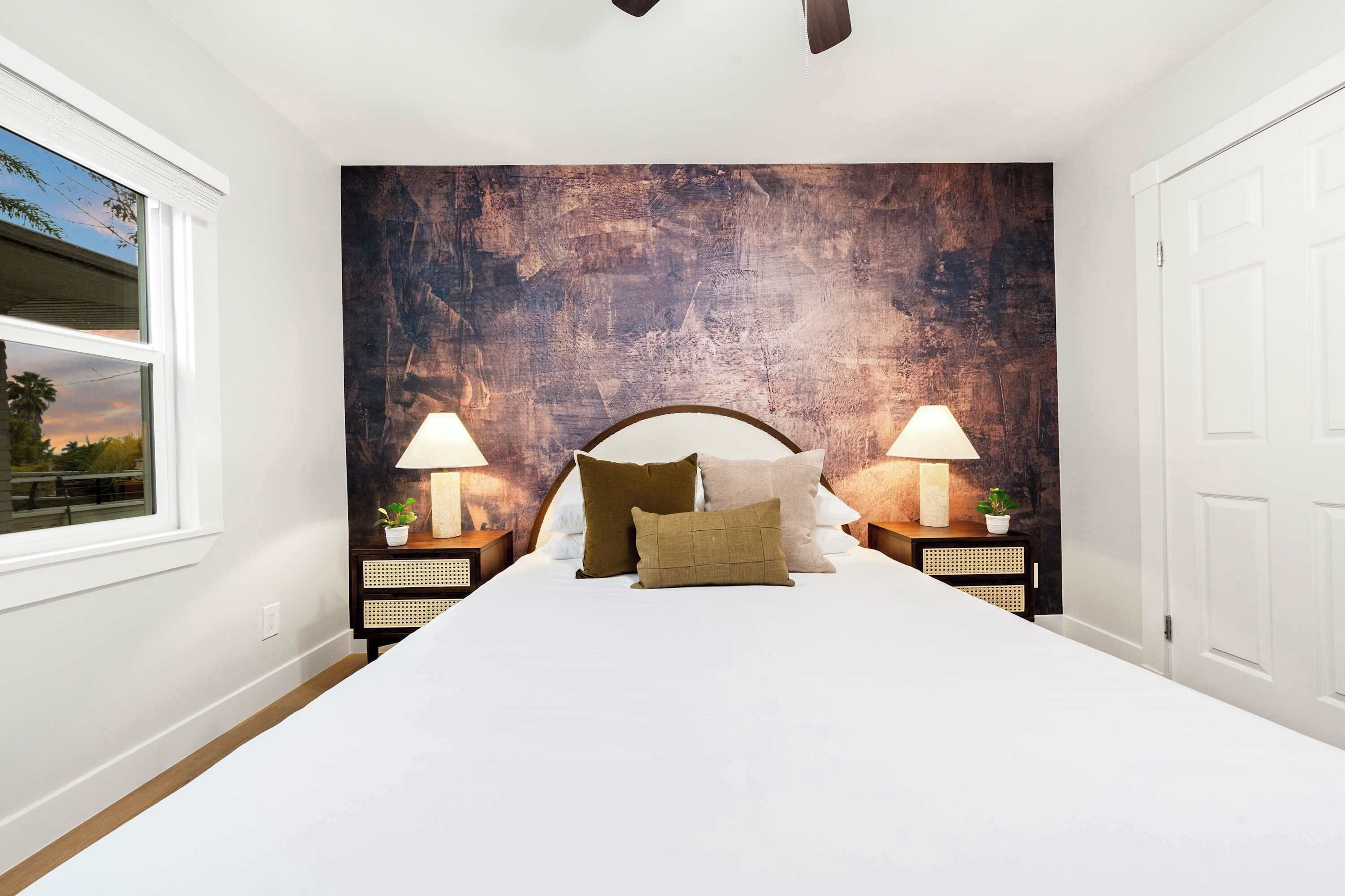 A bedroom with a large bed, two bedside tables with lamps, a textured wall, and a window with a view.