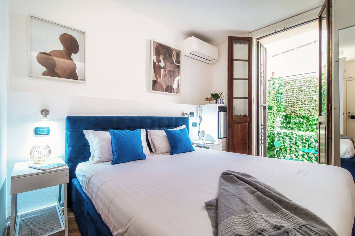 A bedroom with a large bed, a nightstand, a desk, and a window with a view of greenery.