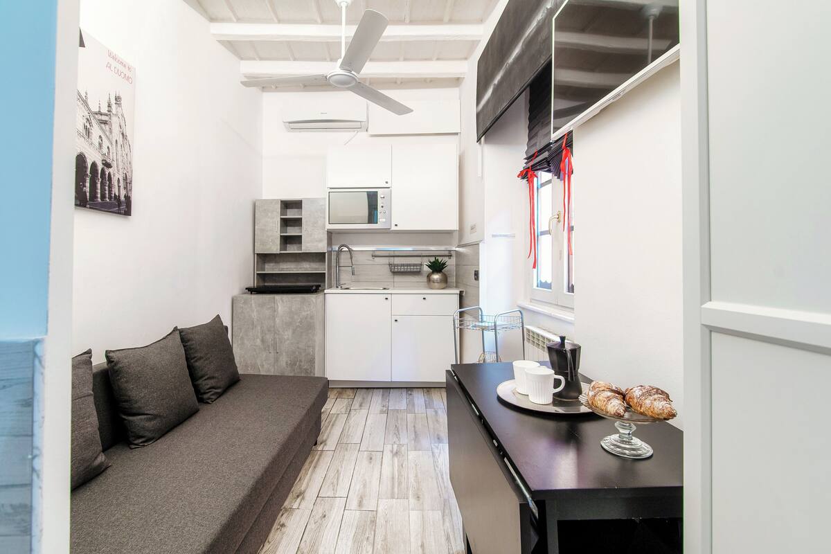 A compact kitchen with a microwave, oven, and sink. There is a sofa with cushions and a small dining table with pastries.