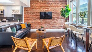 Una sala de estar moderna con pared de ladrillo, televisor de pantalla plana, comedor con taburetes de bar y una cocina al fondo.