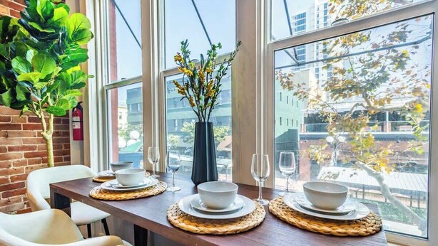 Una mesa de comedor con fuentes blancas y copas de vino, un florero con flores amarillas y vista a edificios a través de amplios ventanales.
