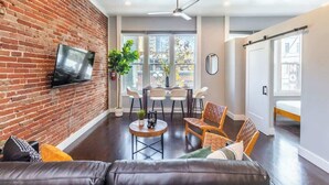 Una sala de estar moderna con pared de ladrillo, televisor de pantalla plana, sofá de cuero, zona de comedor con sillas y un ventanal con vistas a edificios.