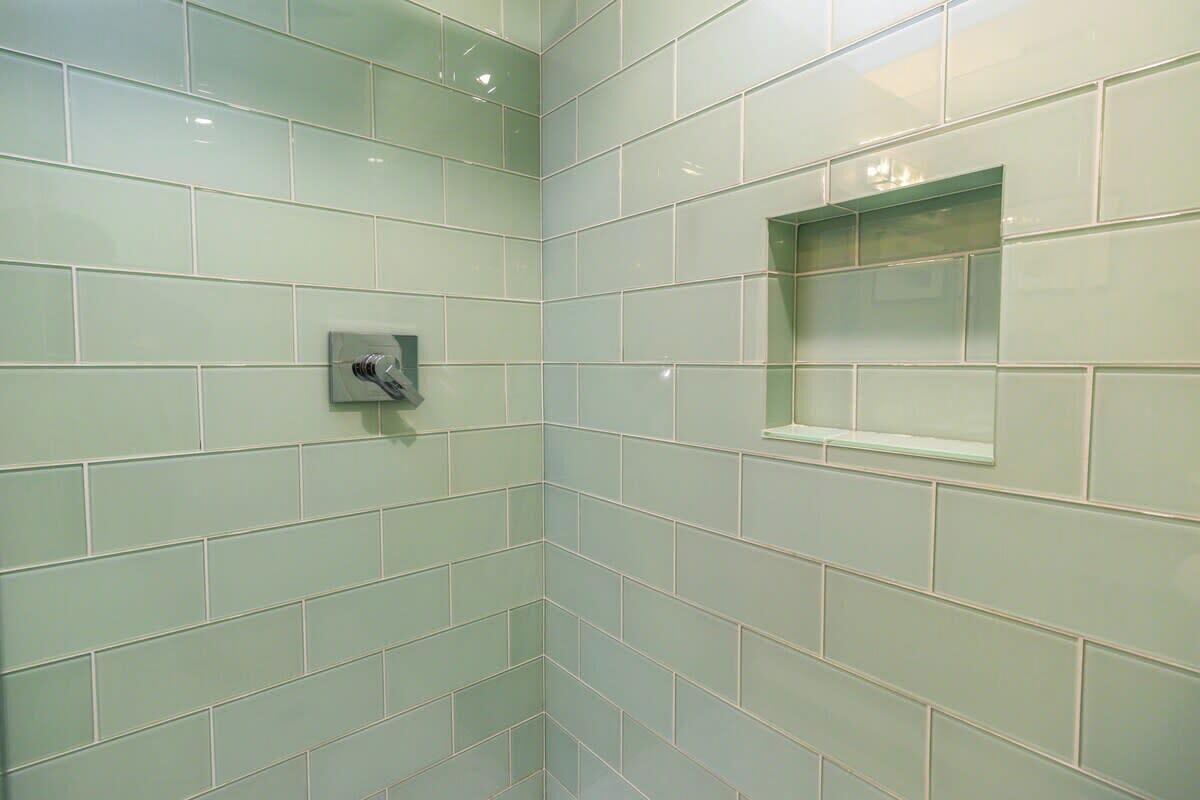 Ein Badezimmer mit hellgrünen Fliesen, einem kleinen Fenster und einer an der Wand montierten Duscharmatur.