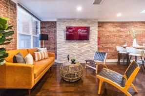 Un espacio lounge moderno con una pared de ladrillo, un televisor de pantalla plana grande montado en la pared, una mesa de centro redonda de mimbre, un sofá amarillo con cojines estampados y una silla de madera con asiento trenzado.