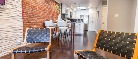 A modern living room with a brick wall, a wooden bench, and two chairs. A kitchen is visible in the background.