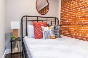 Una cama bien hecha con varios almohadones, una mesita de noche con una lámpara y una pequeña planta. Se ve una pared de ladrillo a la derecha de la cama.