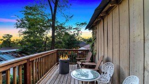 A wooden deck with a glass table and white chairs, a fire pit, and a view of trees and a building in the background. - Escape to Rustic Studio | Fireplace + Modern Comfort (Julian)