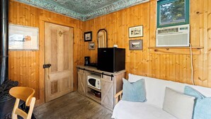 A cozy room with wooden walls, a white sofa, a wooden chair, a black refrigerator, a microwave, and a window with a view of trees. - Escape to Rustic Studio | Fireplace + Modern Comfort (Julian)