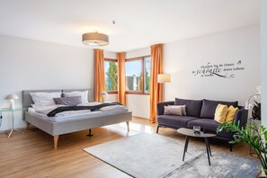 A modern bedroom with a bed, a sofa, a small table, a lamp, and a window with curtains.