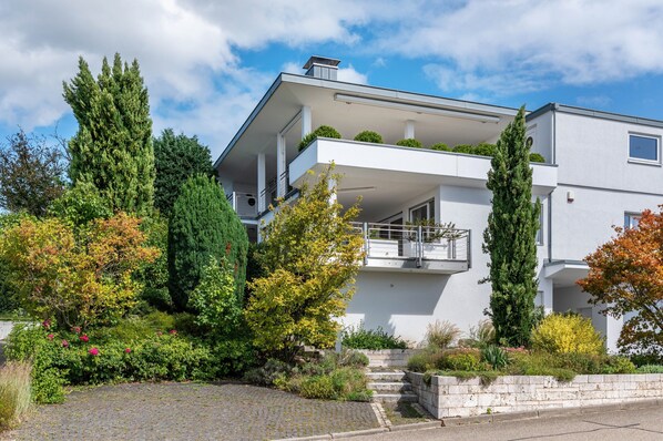 A modern two-story house with a balcony, surrounded by a variety of trees and shrubs.
