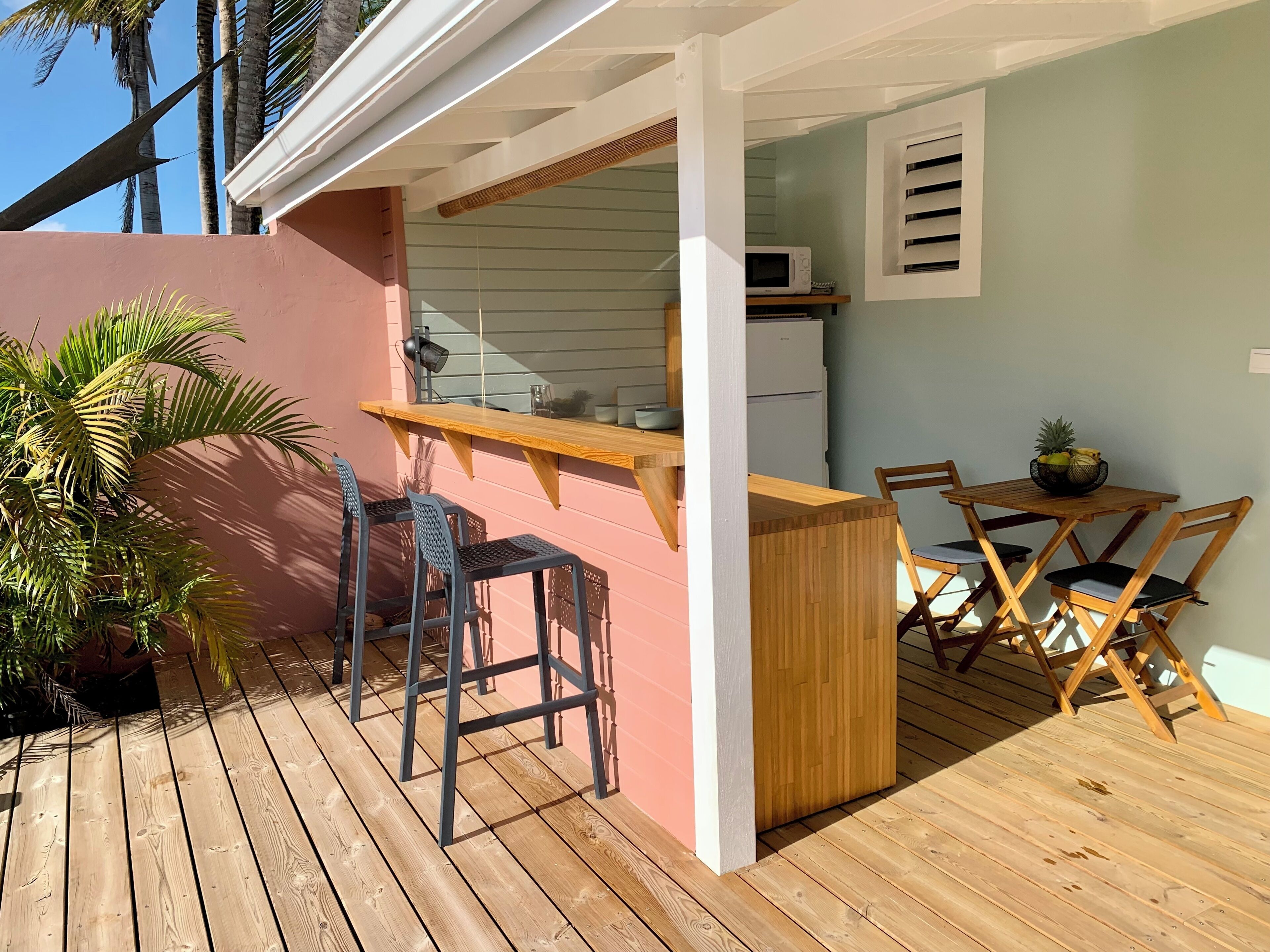 A wooden deck with a kitchen area, including a microwave and a refrigerator. There are two chairs and a table with a fruit bowl.