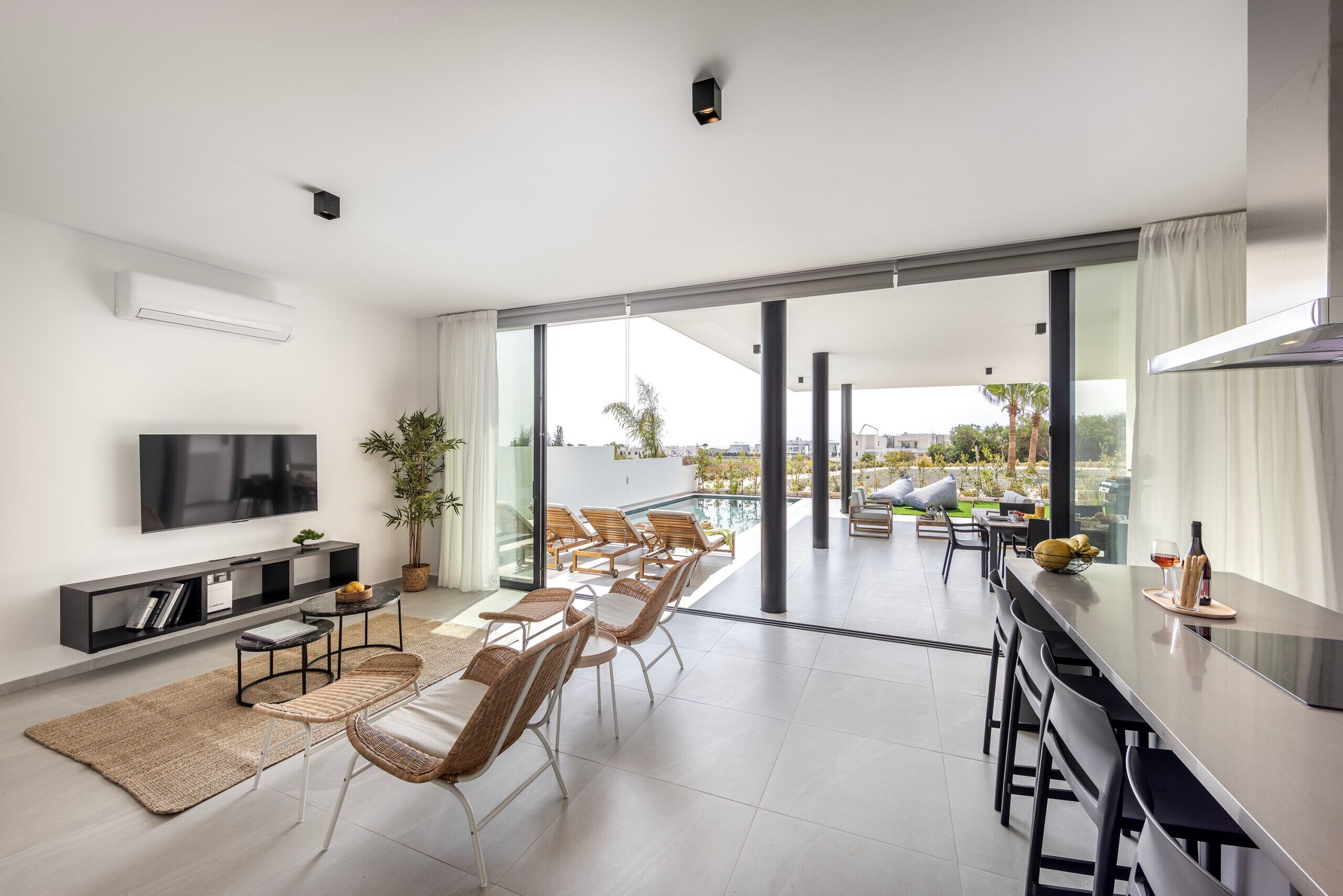 Uma sala de estar moderna com uma TV de tela plana, uma estante de livros, uma área de jantar com cadeiras e uma mesa, e uma grande porta de vidro deslizante que dá para uma varanda com piscina.