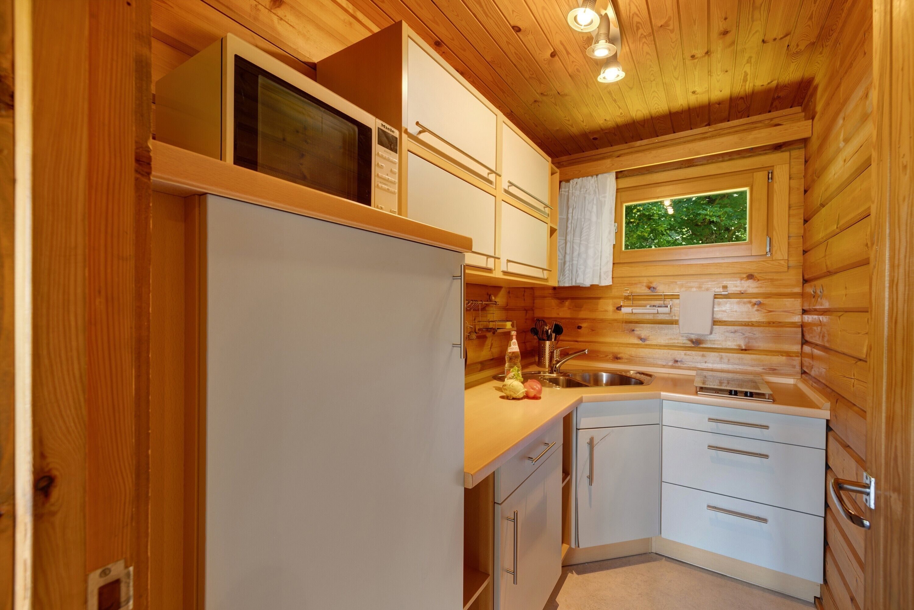 Een kleine keuken met houten wanden, een magnetron, een koelkast, een wastafel en een raam met uitzicht op groen.