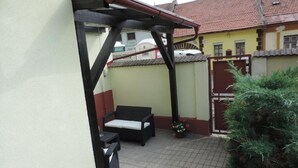 A covered outdoor seating area with a table and chairs, a potted plant, and a building in the background.