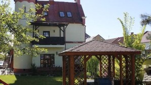 A two-story building with a red roof and a tower-like structure on the left. There is a wooden gazebo in the foreground.