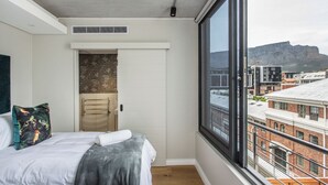 Ein Schlafzimmer mit großem Fenster, einem Bett, einer Tür mit strukturierter Wand und Blick auf Gebäude sowie einen Berg.
