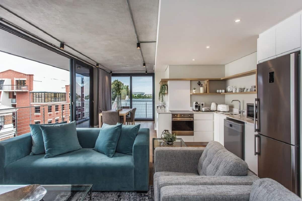 A modern apartment with a living area featuring a teal sofa and a grey sofa, a kitchen with stainless steel appliances, and large windows offering a city view.