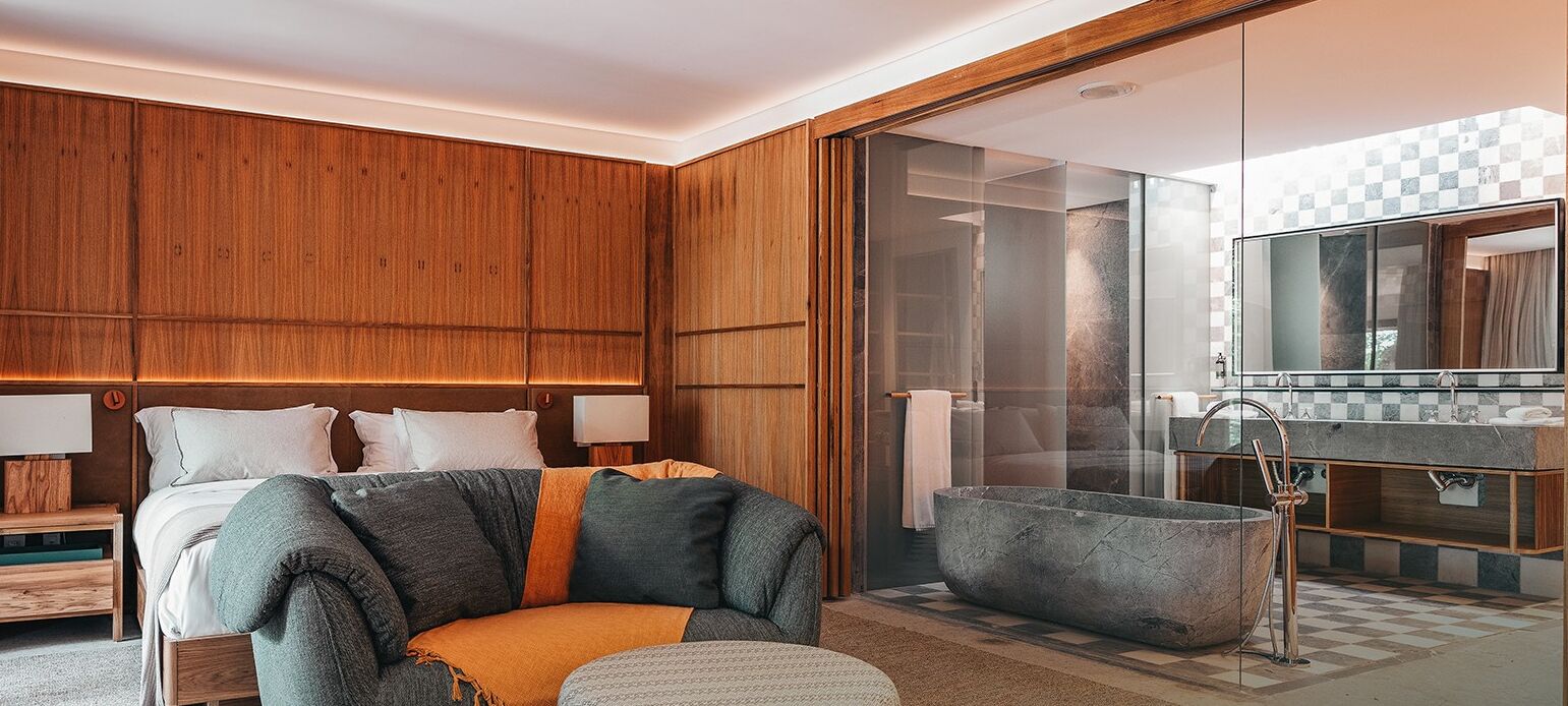 Une chambre d’hôtel moderne avec une tête de lit en bois, un grand lit, un coin salon avec un banc et des coussins, une baignoire en pierre et un grand miroir au-dessus du lavabo.