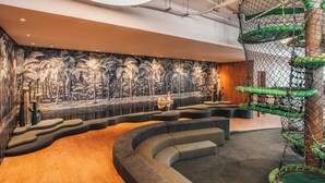 A modern lounge area with a curved seating arrangement, a mural of palm trees, and a vertical play structure. - Clara Arte Resort (Brumadinho)