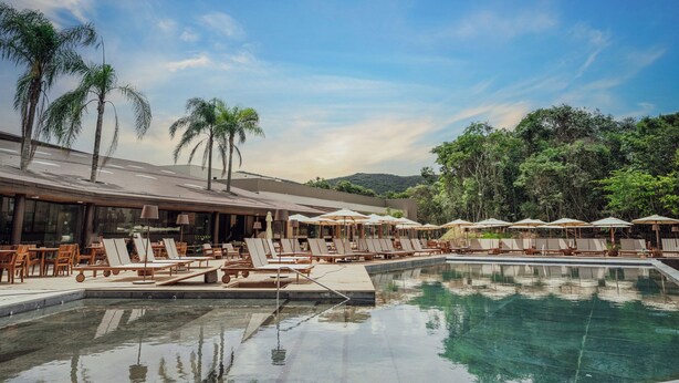 A pool area with lounge chairs and umbrellas, surrounded by palm trees and a building with large windows. - Clara Arte Resort (Brumadinho)