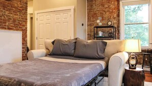 A bedroom with a bed, a nightstand, a lamp, a clock, and a brick wall. - NULU/Butchertown The Sidecar Suite Butchertown Salon/Spa (Louisville)