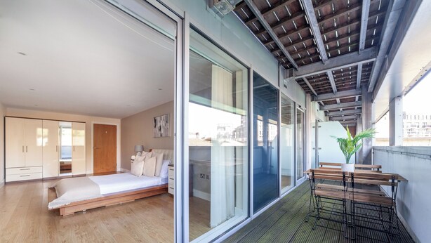 A modern bedroom with a large bed, sliding glass doors, and a balcony with a wooden bench. - Cosy 1 Bed Flat in Greenwich (London)