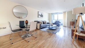 A modern living room with a sofa, coffee table, and chairs. There is a large mirror on the wall and a lamp on a tripod stand. - Cosy 1 Bed Flat in Greenwich (London)