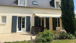 Een huis met twee verdiepingen, voorzien van een pannendak, een overdekt terras met tuinmeubilair en een tuin met bloemen en een hoge boom.