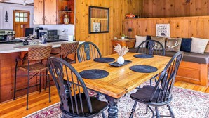 A wooden interior with a dining table, chairs, a kitchen area, and a living space with a sofa and cushions.