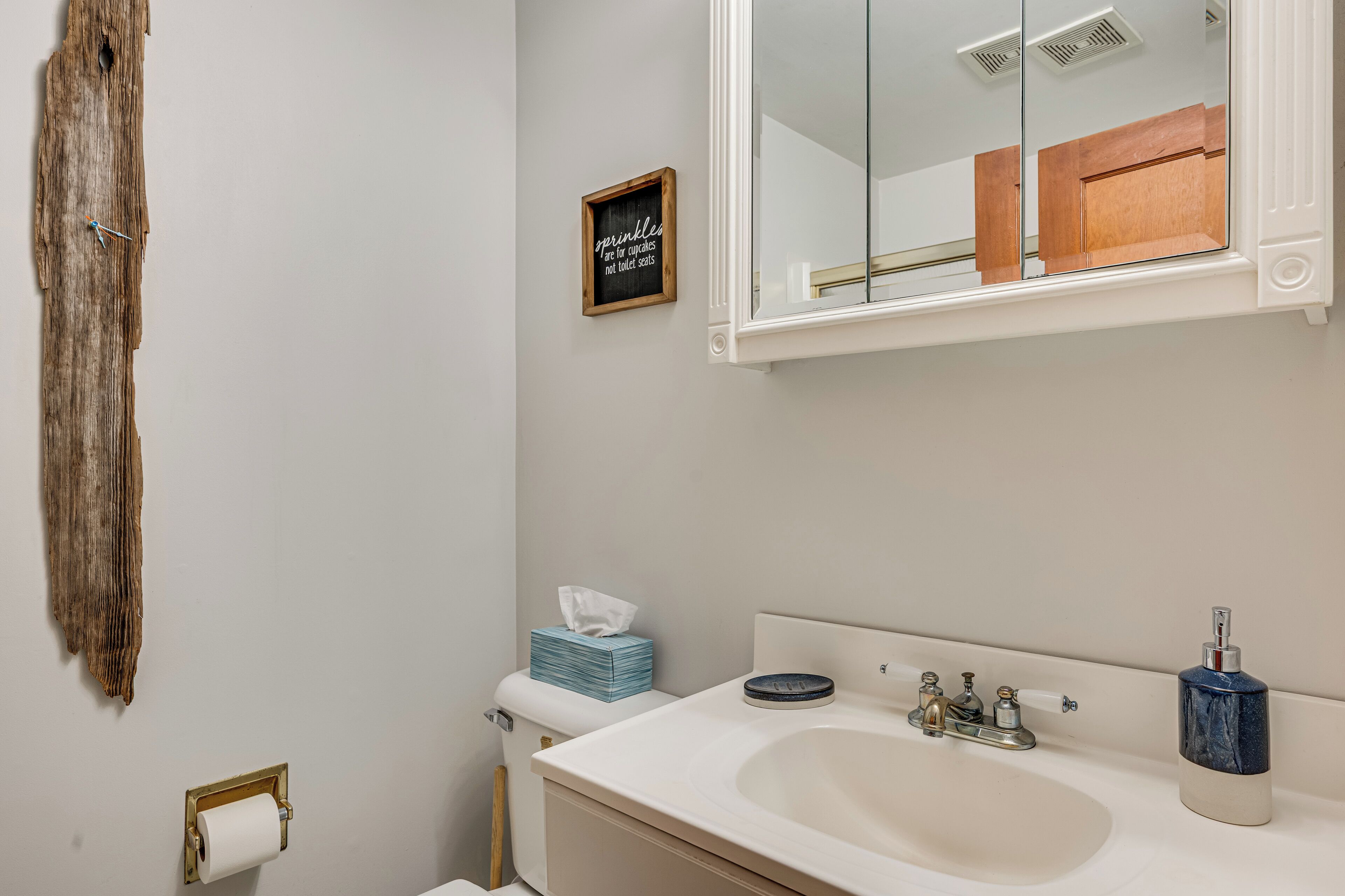 Une salle de bain avec un lavabo, un miroir et des toilettes. Un panneau en bois est accroché au mur, et un rouleau de papier toilette est également fixé au mur.