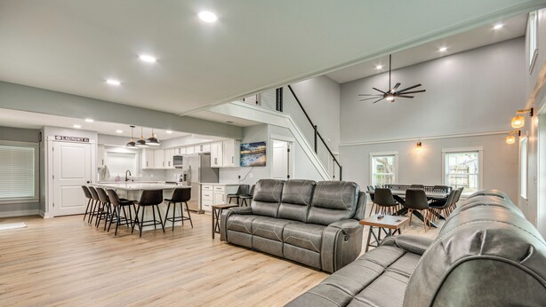 A spacious living area with a grey leather sofa, a dining table with chairs, a kitchen area, and a staircase leading to an upper level.