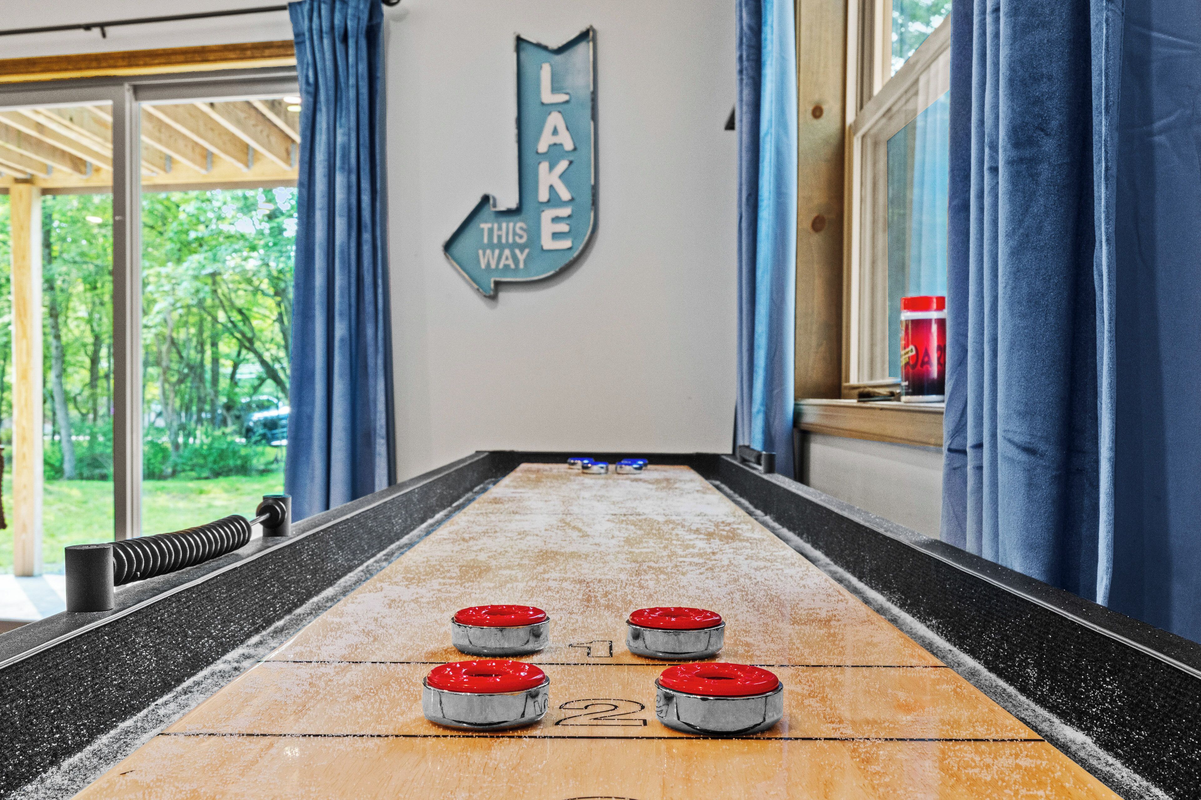A shuffleboard table with four pucks, a sign reading "LAKE THIS WAY", and a view of trees through large windows.