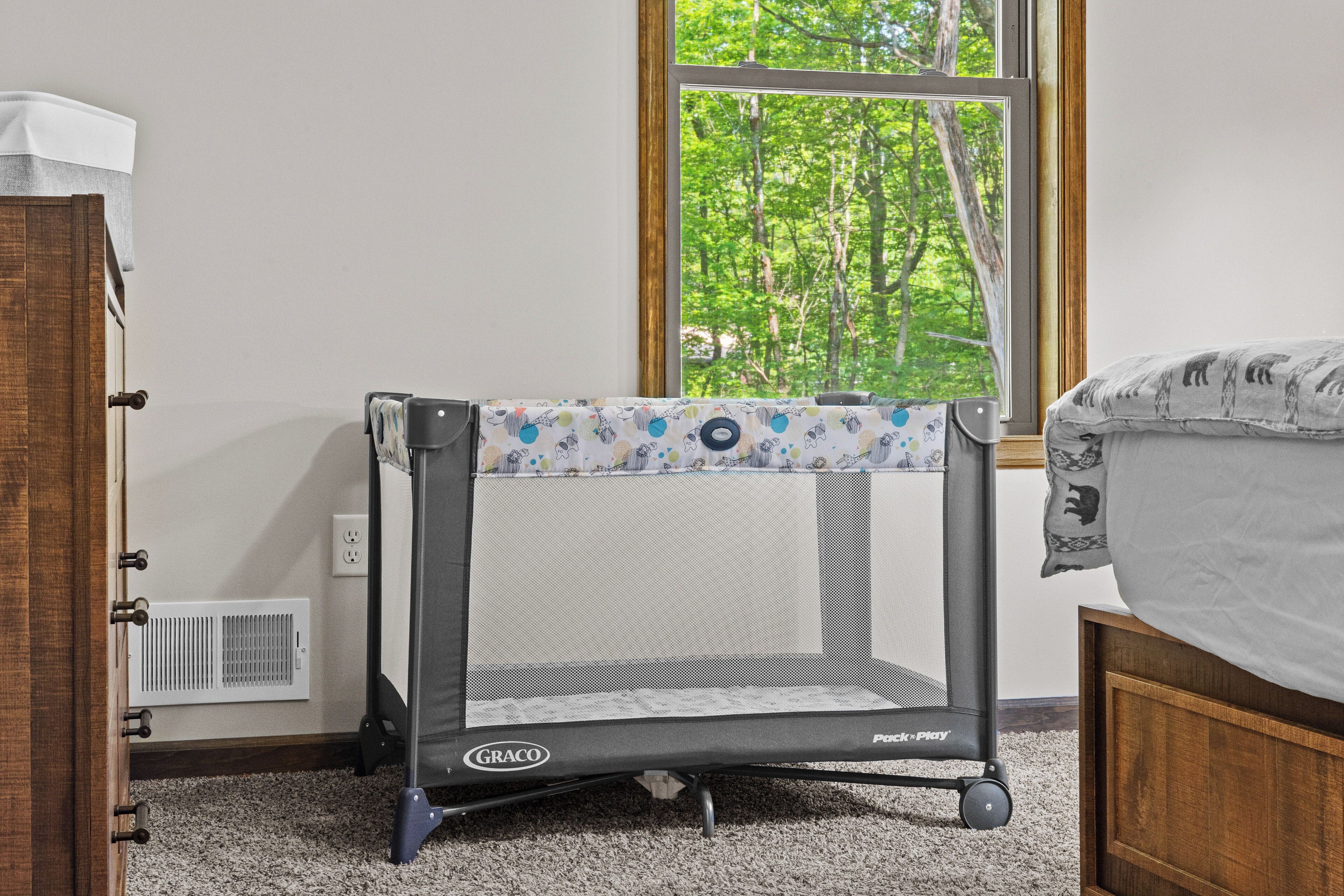 Una habitación con una parque infantil Graco cerca de una ventana que da a árboles.