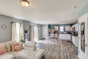 Una sala moderna con un sofá blanco, un comedor con una mesa y sillas blancas, y una cocina con armarios y electrodomésticos blancos.