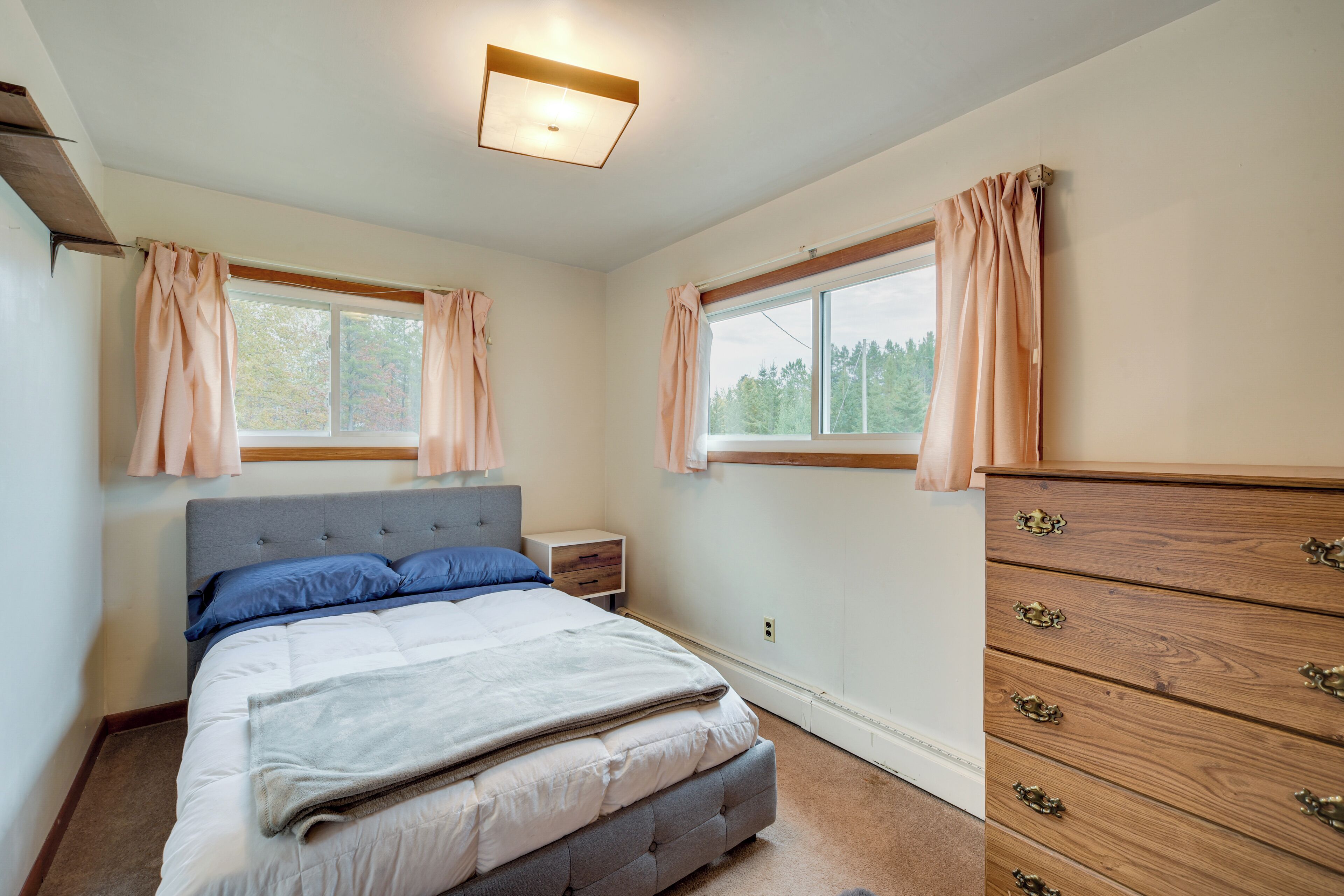 A bedroom with a bed, nightstand, dresser, and two windows with curtains.
