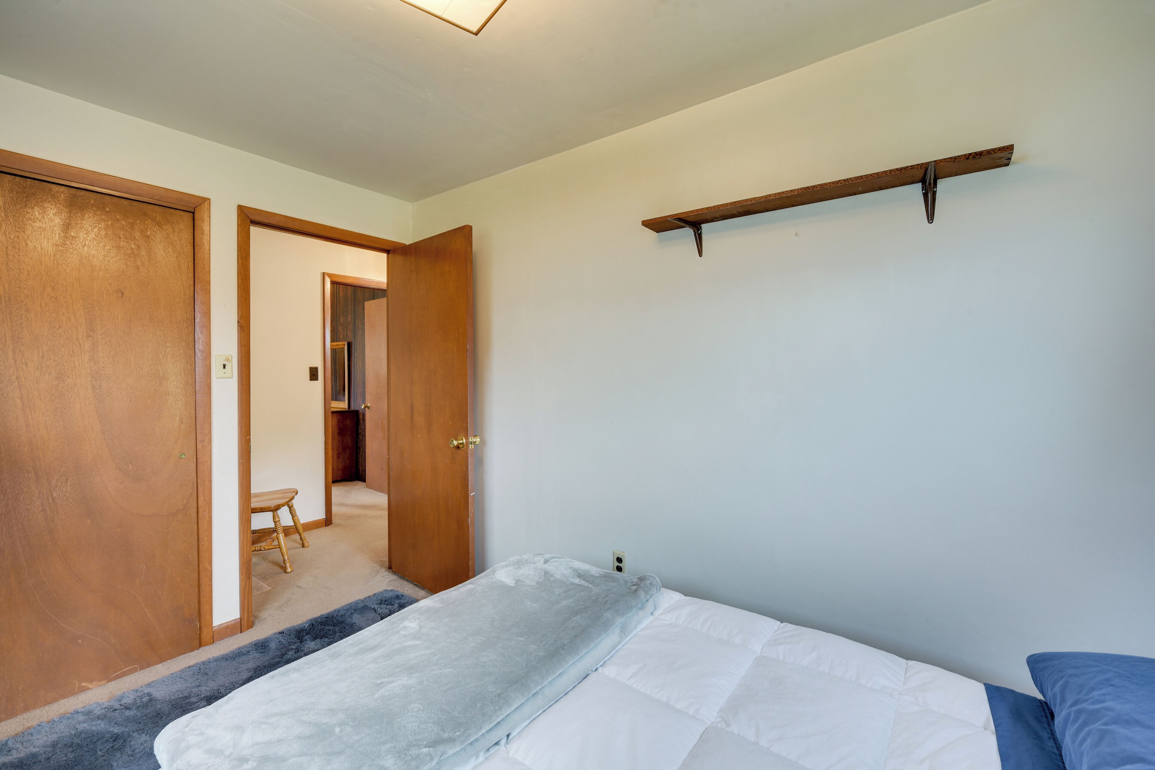 Un dormitorio con cama, una puerta de madera, un taburete pequeño y una estantería en la pared.