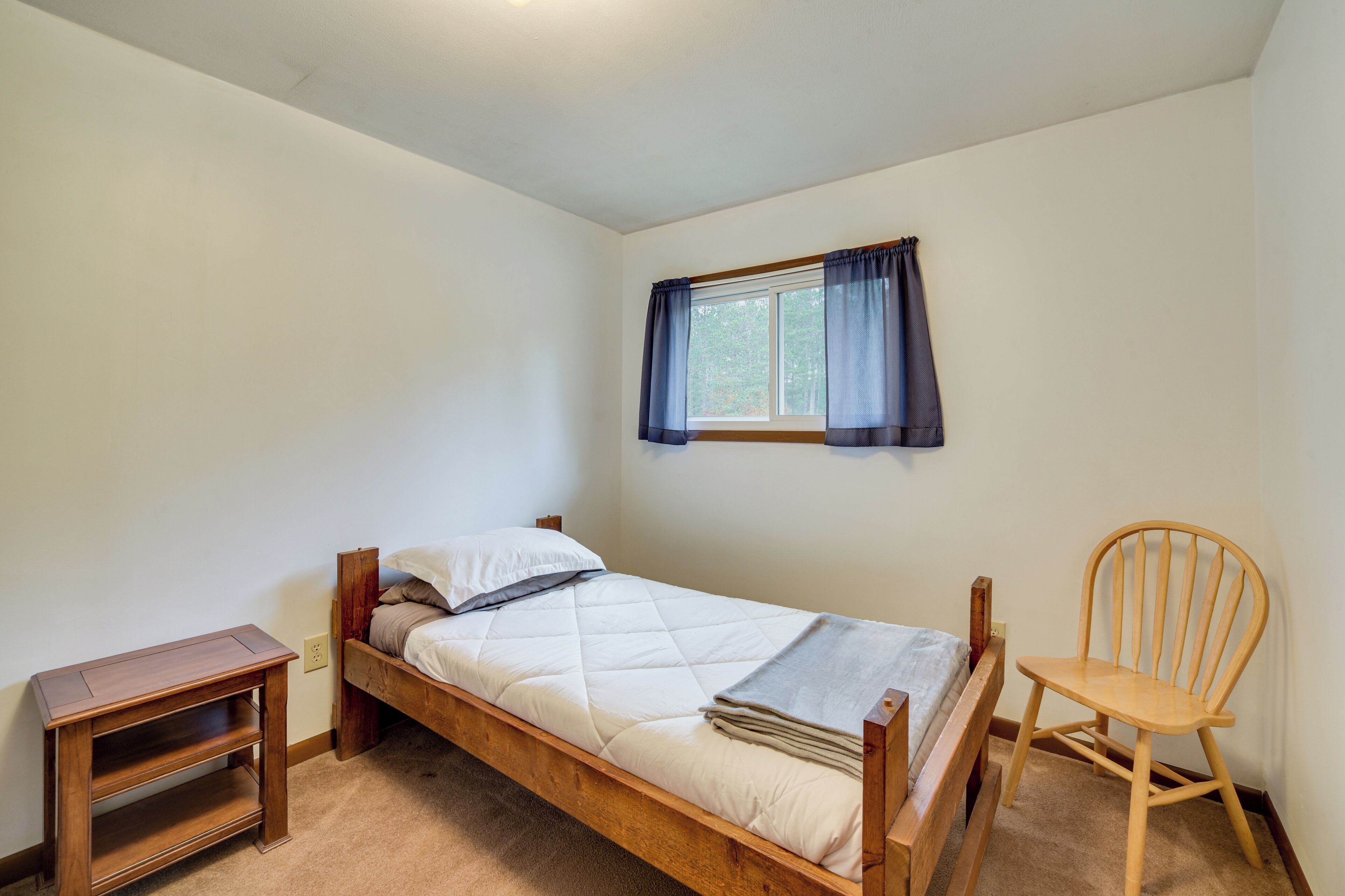A single bed with a wooden frame, a nightstand, a chair, and a window with curtains.
