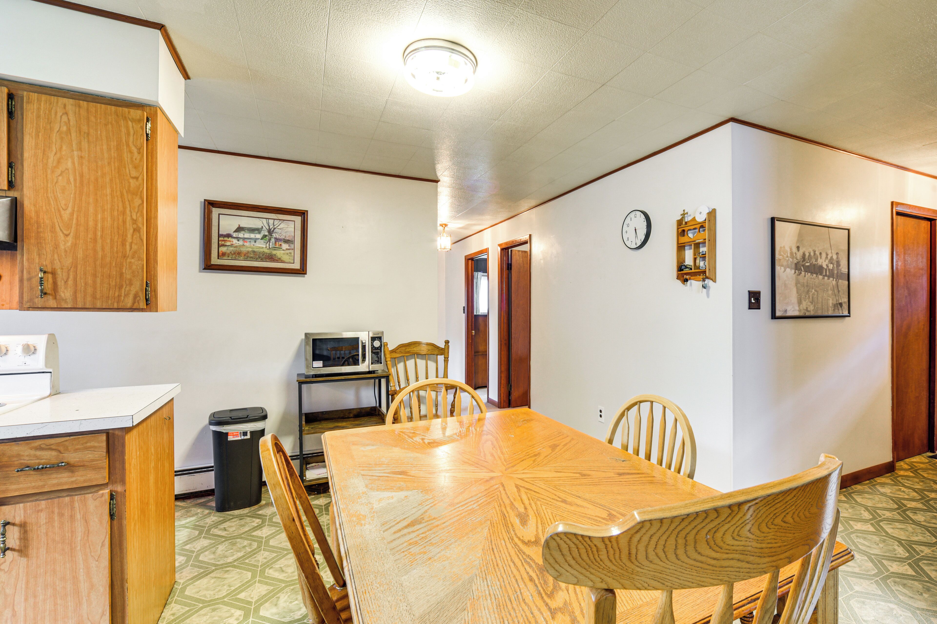 A dining area with a wooden table and chairs, a microwave, a trash can, and a clock on the wall.