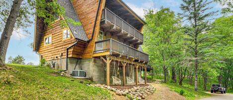 A wooden cabin with a balcony and a covered porch, surrounded by trees and a road.