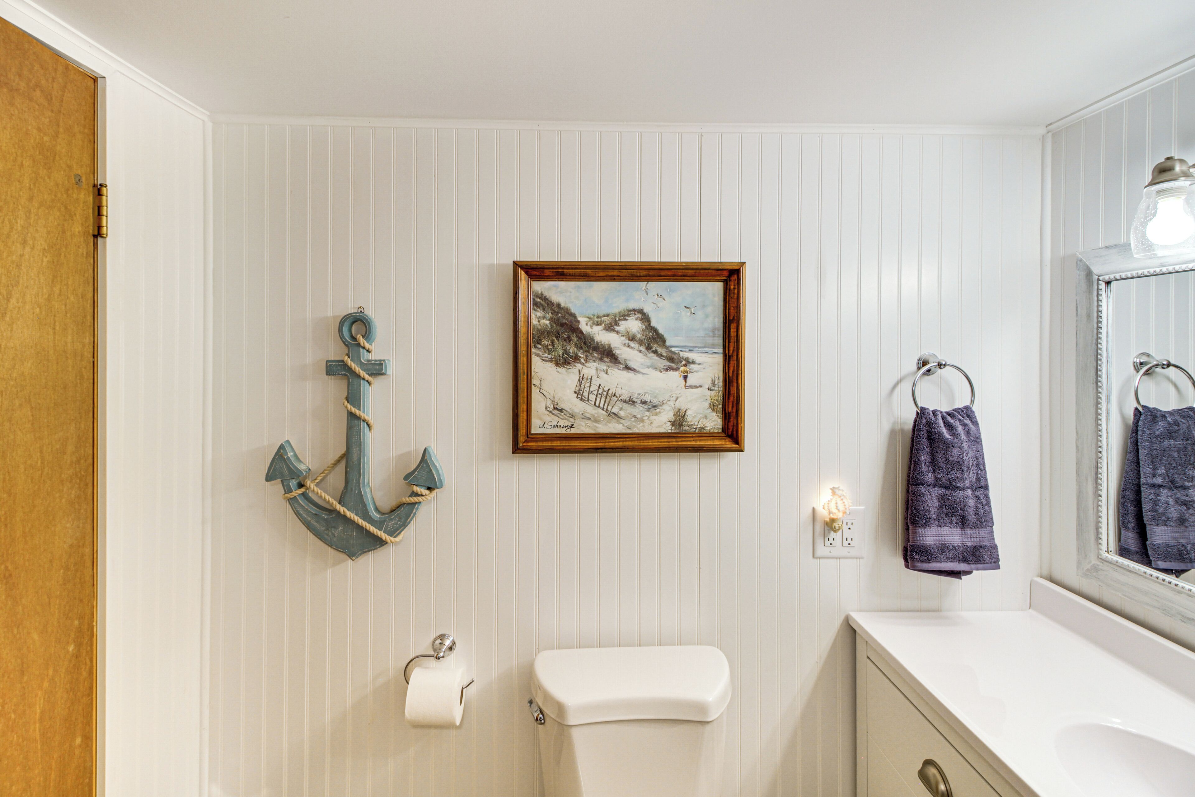 A bathroom with a striped wall, a framed painting, an anchor decoration, a toilet, and a sink with a towel hanging on the wall.