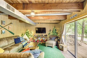 Interior acogedor de una cabaña con techo de madera, televisor de pantalla plana, chimenea y un ventanal con vistas a un bosque.